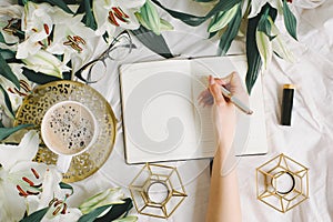 Woman writing notes in her notebook while lying on the white bed with coffee cup, flowers and glasses. Flat lay style
