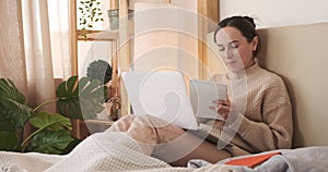 Woman writing notes looking at laptop in bed