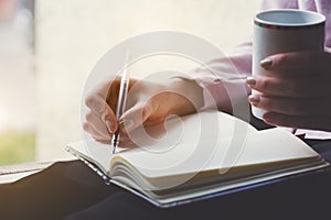 Woman writing notes on notebook with pen and drinking coffee