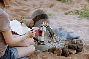 a woman& x27;s hand holds a red cup of coffee or tea against the background of a campfire on a hike. hiking and outdoor