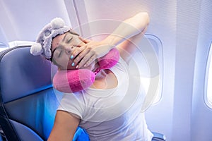 Woman yawns while sitting on the plane with a mask and a pillow for sleeping.