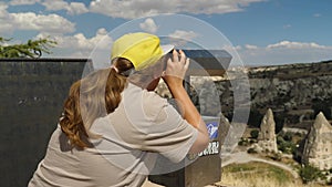 A woman in a yellow baseball cap looks out over the mountains through sightseeing binoculars. Sightseeing through