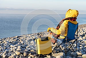 Woman in yellow coat reaching the destination and sitting on beach chairs on the seaside at sunrise. British cold