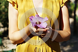 Woman in a yellow dress holding a fresh purple hibiscus flower in a park