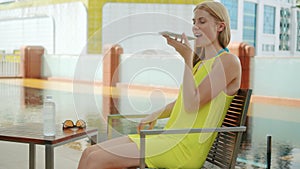 Woman in yellow dress sitting in chair near swimming pool. Using her smartphone, talking with friend