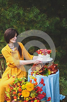 Woman in yellow dress on wooden chair by round table with blue tablecloth, eating white cake with berries, maple leaves, apples,