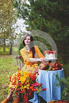 Woman in yellow dress on wooden chair by round table with blue tablecloth, eating white cake with berries, maple leaves, apples,