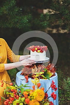 Woman in yellow dress on wooden chair by round table with blue tablecloth, eating white cake with berries, maple leaves, apples,