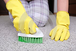 Woman in yellow gloves with a green brush cleaning and brushing carpet, removing stains and wool from it and doing