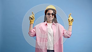 Woman in yellow helmet and gloves pointing and gesturing against blue studio background
