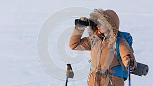 Woman in yellow hooded down jacket with a backpack and ski poles looks through binoculars standing on a snowy plain in