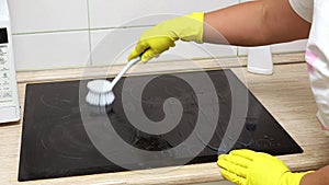 Woman in yellow household gloves rub detergent in circular motion over surface of electric ceramic hob in kitchen