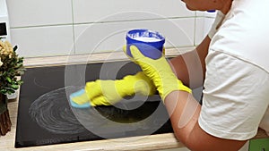 Woman in yellow household gloves soak sponge in cleaner then scrub surface of electric ceramic hob in kitchen. Housewife