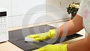 Woman in yellow household gloves sponge surface of electric ceramic hob in kitchen. Housewife carry out wet cleaning of