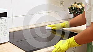 Woman in yellow household gloves sponge surface of electric ceramic hob in kitchen. Housewife carry out wet cleaning of