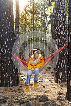 Woman with yellow jacket sit down and rest on a colorful hammock on the forest woods enjoying outdoor leisure activity and