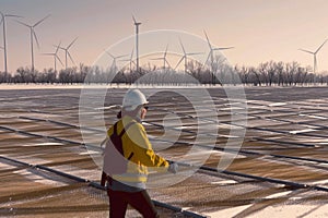 A woman in a yellow jacket is walking on a field with a bunch of wind turbines i