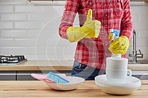 Woman in yellow protective glove with plates, dishes, and cloth showing ok sign with thumb up. Kitchen background