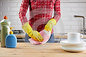 Woman in yellow protective glove with plates, dishes, plastic bottles and cloth, wooden table on kitchen background