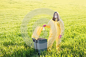 A woman in a yellow protective suit stands in the middle of a green field and holds a plastic bottle in her hands to throw in the