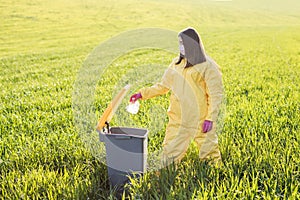 A woman in a yellow protective suit stands in the middle of a green field and holds a plastic bottle in her hands to throw in the