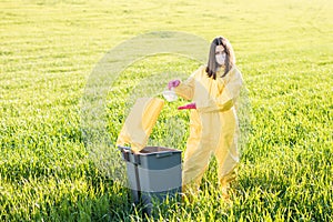 A woman in a yellow protective suit stands in the middle of a green field and holds a plastic bottle in her hands to throw in the