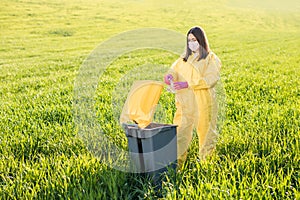 A woman in a yellow protective suit stands in the middle of a green field and holds a plastic bottle in her hands to throw in the