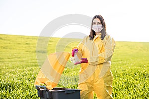 A woman in a yellow protective suit stands in the middle of a green field and holds a plastic bottle in her hands to throw in the