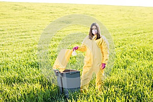 A woman in a yellow protective suit stands in the middle of a green field and holds a plastic bottle in her hands to throw in the