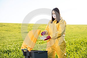 A woman in a yellow protective suit stands in the middle of a green field and holds a plastic bottle in her hands to throw in the