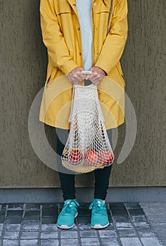 Woman in a yellow raincoat carrying string bag with fresh bananas and apples on the street