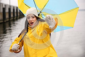 Woman in yellow raincoat with umbrella caught in gust of wind near river