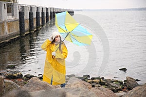 Woman in yellow raincoat with umbrella caught in gust of wind near river