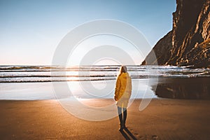 Woman in yellow raincoat walking alone on beach enjoying sunset ocean view