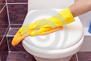 Woman in yellow rubber gloves cleaning toilet seat cover with orange cloth. Bathroom and toilet hygiene.