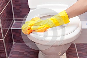 Woman in yellow rubber gloves cleaning toilet seat with orange cloth. Bathroom and toilet hygiene.