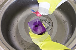 Woman in yellow rubber gloves pours washing solution on a sponge , a sink on background.
