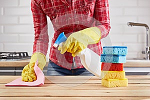 Woman in yellow rubber protective gloves cleaning wooden table with spray, sponge and cloth, kitchen background