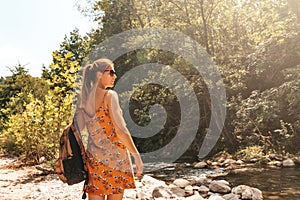 Woman in yellow sundress enjoying a walk near river in national park. Solo traveler outdoors with backpack, view from