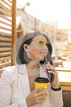 Woman in yellow sunglasses with earbuds in ears with phone, glass of coffee looks up smiling.