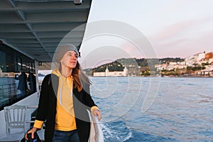 Woman in a yellow sweater and gray beret standing on deck of cruise ship and looking at landscape. Sunset light, golden hour.