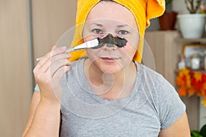 A woman in a yellow towel on her head and in a gray T-shirt applies a mask of blue clay to her face with a brush