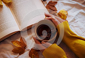 A woman in yellow warm sweater relaxing reading a book holding hot mug of aroma spice tea on the table with yellow autumn leaf . C