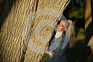 Woman, young, pretty, blonde, with blue eyes and denim dress, playing hide and seek behind the trunk of a large tree receiving the