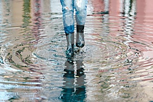Womans feet with black rubber boots and blue jeans standing in a puddle of water after rain on a city street. Front view