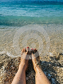 Womans feet with red nails pedicure and sand on golden sandy beach in blue turquoise sea waters. Holiday concept near