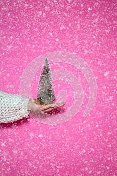 Womans hand holding a christmas tree on pink background with snow