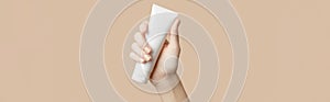 A womans hand holding a white cosmetic tube on a beige background. Care products