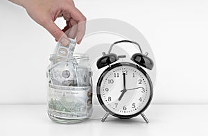 Womans hand put dollar bancnote into a glass jar with alarm clock