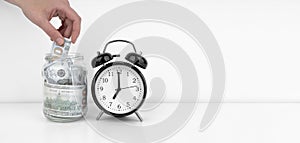 Womans hand put dollar bancnote into a glass jar with alarm clock
